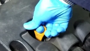 Desnom rukom u zaštitnoj plavoj rukavici obuhvata kažiprstom prstenastu žutu dršku merača nivoa ulja u motoru na gornjem tamno sivom zaštitnom poklopcu motora. Izvlačenjem i brisanjem merača se obavlja priprema za proveru nivoa i kvaliteta ulja u motoru.