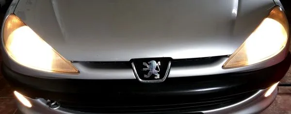 Slika predstavlja pregled ispravnosti prednjih farova vozila u postupku Provera osvetljenja vozila. Na slici je prednji deo sivog Peugeot vozila na kome su upaljeni farovi (pozicija i dugo svetlo) i prednji farovi za maglu u donjem delu prednje maske ispod crnog branika.