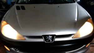 Slika predstavlja pregled prednjeg spoljašnjeg osvetljenja vozila u postupku Provera osvetljenja vozila. Na slici je prednji deo sivog Peugeot vozila sa upaljenim prednjim farovima i svetlima za maglu koji se nalaze u donjoj sivoj maski ispod crnog branika.