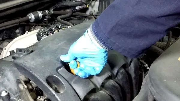 Slika predstavlja postupak Tehničko održavanje i kontrolni pregled vozila. Serviser u zaštitnoj plavoj rukavisi desnom rukom je obuhvatio žuti držač merne šipke ulja motora. na slici se vidi tamno sivi poklopac motora u motornom prostoru vozila.