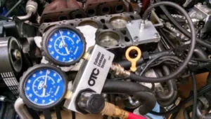 Slika predstavlja link ka stranici test curenja cilindra u okviru teme Ispitivanje mehanike motora – procena stanja. Na slici je prikazan dvostruki plavi manometar koji su povezani na motor u pozadini.